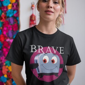 May include: Black t-shirt featuring a cartoon toaster character with the word "BRAVE" above it. The toaster has a smiling face, red eyes, and is set against a pink circle. The shirt is worn by a person.
