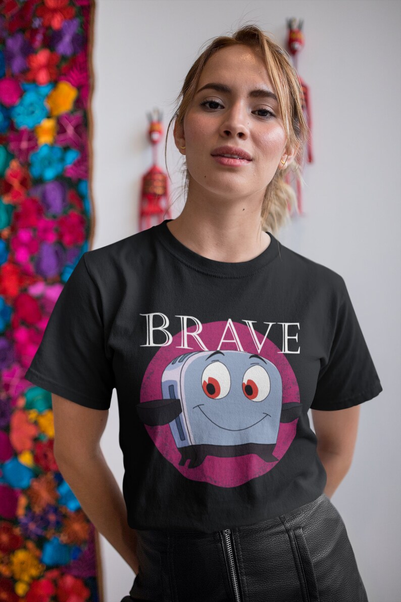 May include: Black t-shirt featuring a cartoon toaster character with the word "BRAVE" above it. The toaster has a smiling face, red eyes, and is set against a pink circle. The shirt is worn by a person.