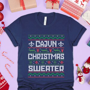 May include: A navy blue t-shirt with a white, red, and green Christmas sweater design. The design features the text "Cajun Christmas Sweater" and images of alligators and crawfish.