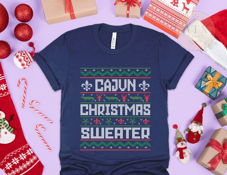 May include: A navy blue t-shirt with a white, red, and green Christmas sweater design. The design features the text "Cajun Christmas Sweater" and images of alligators and crawfish.