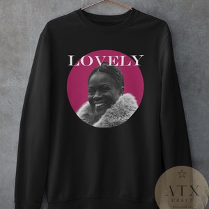 May include: Black sweatshirt with a pink circle featuring a woman smiling. The text "LOVELY" is above the circle. The sweatshirt has a white star and the text "ATX CRAFT designed in austin, tx" on a brown tag.