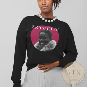 May include: Black sweatshirt with a pink circle and a black and white photo of a woman smiling. The text "LOVELY" is printed in white above the circle.