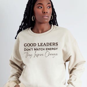 May include: A beige sweatshirt with the text "GOOD LEADERS DON'T MATCH ENERGY They Inspire Change" printed in brown. The text is in a bold, sans-serif font. The sweatshirt is being modeled by a person with dark skin and long, dark hair.