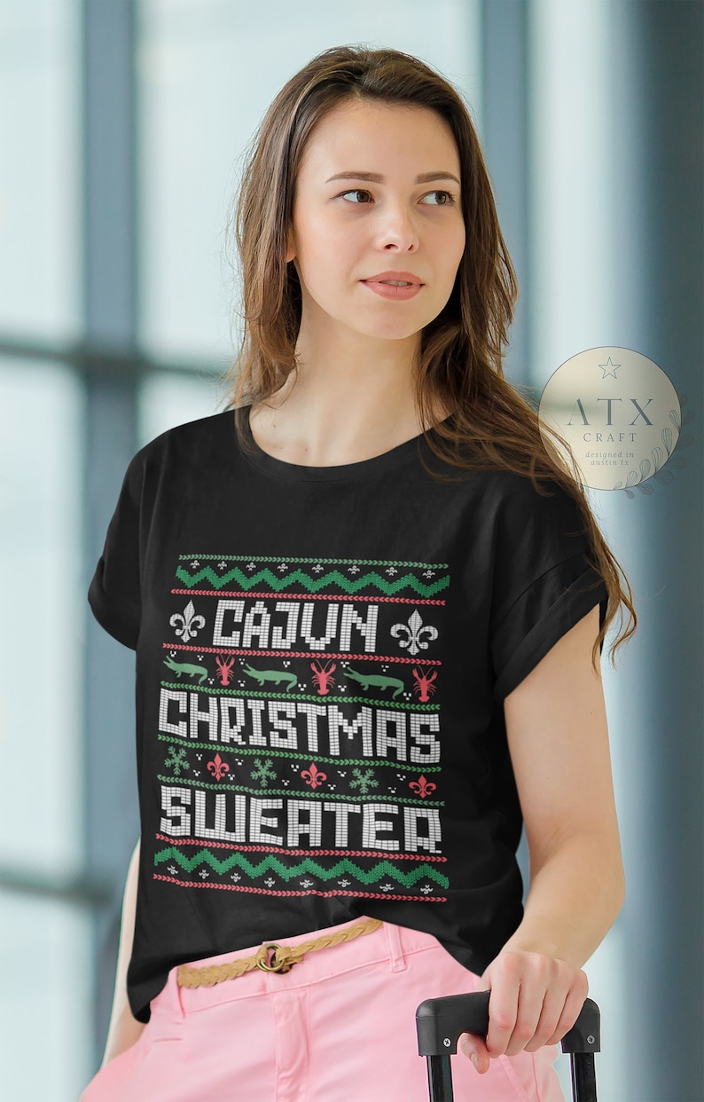 May include: A black t-shirt with a festive design featuring the text "Cajun Christmas Sweater" in white. The design includes red and green snowflakes, fleur-de-lis, alligators, and crawfish.