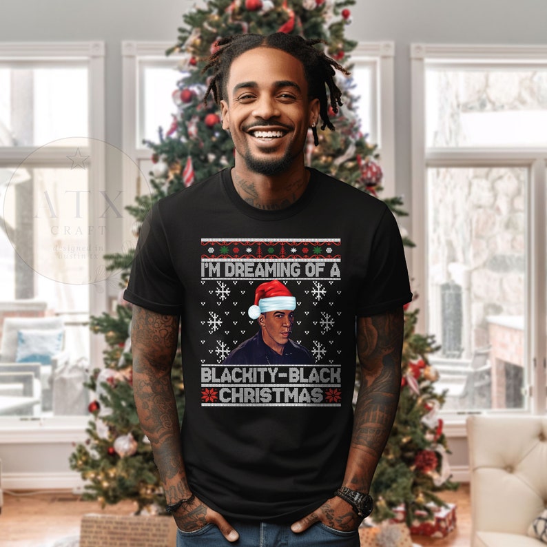 Op de afbeelding: Zwarte man die een zwart T-shirt draagt met een kersttrui-ontwerp. Het ontwerp toont een man met een kerstmanmuts en de tekst "I'm Dreaming of a Blackity-Black Christmas".