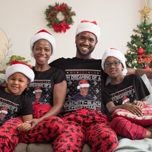 Op de afbeelding: Een gezin van vier personen draagt bijpassende zwarte kersttruien met een rood en wit sneeuwvlokkenpatroon. De truien bevatten een portret van een man met een kerstmanmuts en de tekst "I'm Dreaming of a Blachity-Black Christmas". Het gezin zit op een bank voor een kerstboom.