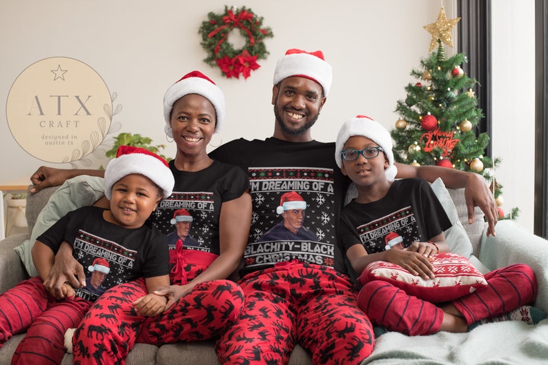 Op de afbeelding: Een gezin van vier personen draagt bijpassende zwarte kersttruien met een rood en wit sneeuwvlokkenpatroon. De truien bevatten een portret van een man met een kerstmanmuts en de tekst "I'm Dreaming of a Blachity-Black Christmas". Het gezin zit op een bank voor een kerstboom.