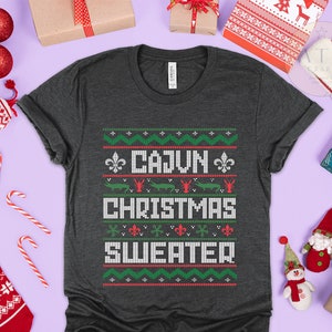 May include: A dark gray t-shirt with a festive Christmas sweater design. The design features a green, red, and white pattern with the text "Cajun Christmas Sweater".