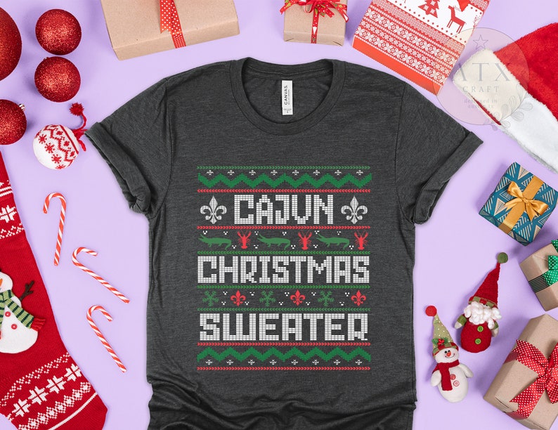 May include: A dark gray t-shirt with a festive Christmas sweater design. The design features a green, red, and white pattern with the text "Cajun Christmas Sweater".