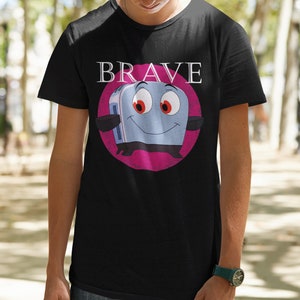 May include: Black t-shirt featuring a cartoon toaster character with red eyes and a smiling face, set against a pink circle. The word "BRAVE" is printed above the toaster in white letters. The shirt is made of a soft material.
