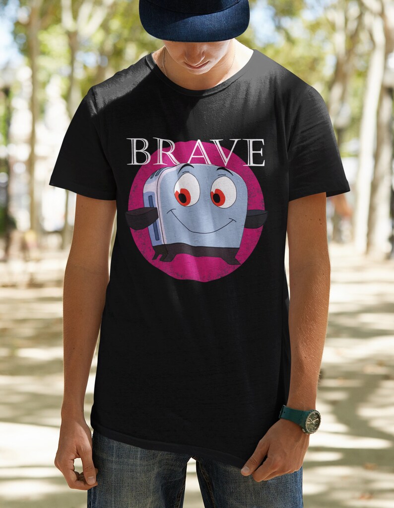 May include: Black t-shirt featuring a cartoon toaster character with red eyes and a smiling face, set against a pink circle. The word "BRAVE" is printed above the toaster in white letters. The shirt is made of a soft material.