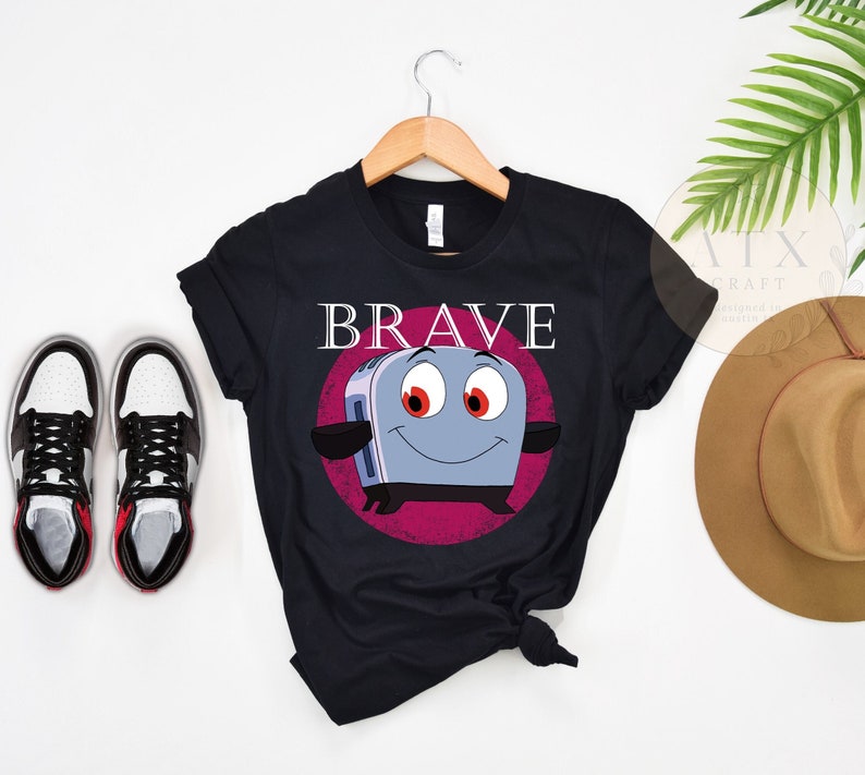 May include: Black t-shirt featuring a cartoon toaster character with red eyes and a smiling face, set against a pink circle. The word "BRAVE" is printed above the toaster. The shirt is displayed on a wooden hanger.