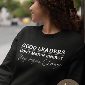 May include: Black sweatshirt with white text that reads "Good Leaders Don't Match Energy They Inspire Change". The bottom right corner of the sweatshirt has a white star and the text "ATX Craft designed in austin tx".