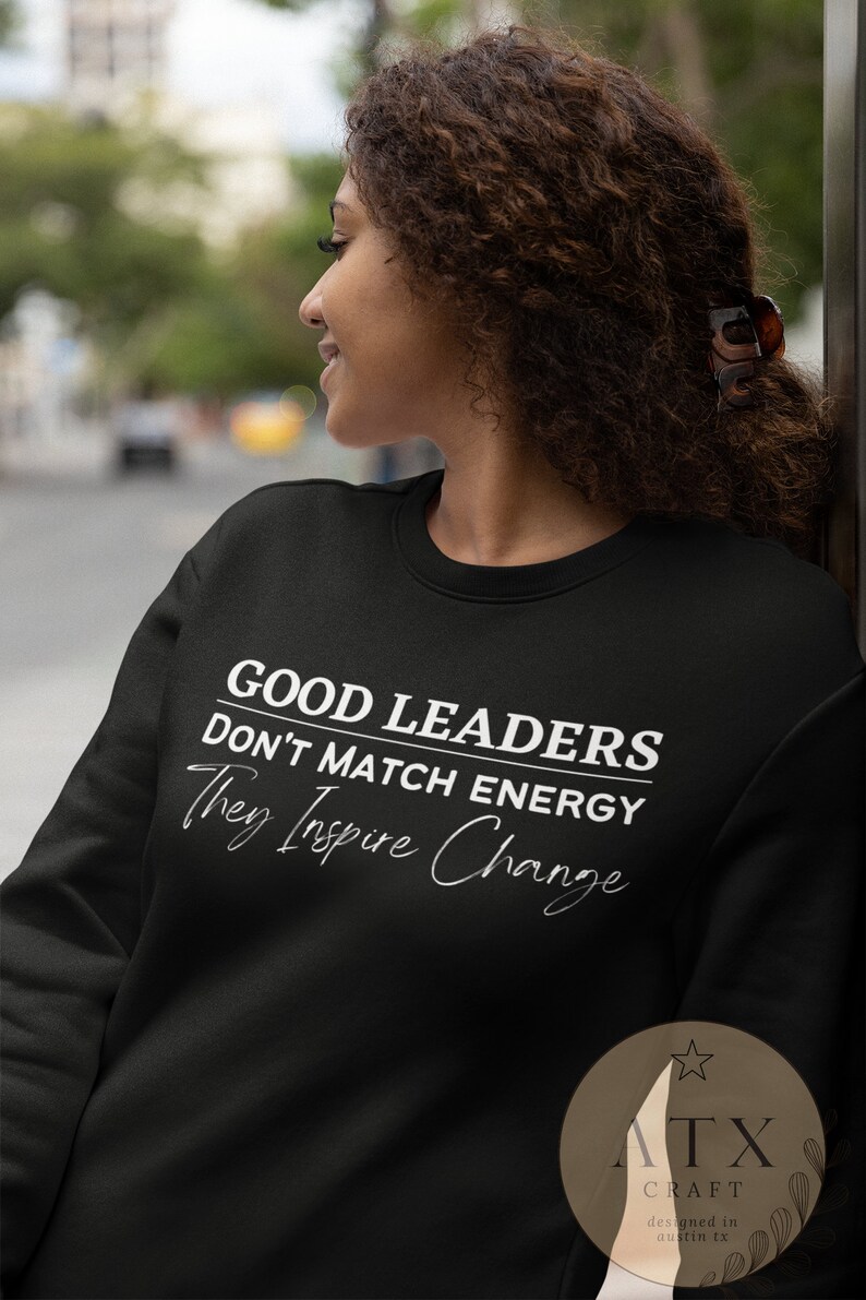 May include: Black sweatshirt with white text that reads "Good Leaders Don't Match Energy They Inspire Change". The bottom right corner of the sweatshirt has a white star and the text "ATX Craft designed in austin tx".