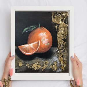 Puede incluir: Pintura de una naranja en un marco blanco. La obra de arte representa una naranja entera con una hoja verde y una rodaja de naranja. Acentos de pan de oro adornan el fondo oscuro. La obra de arte es sostenida por una persona.