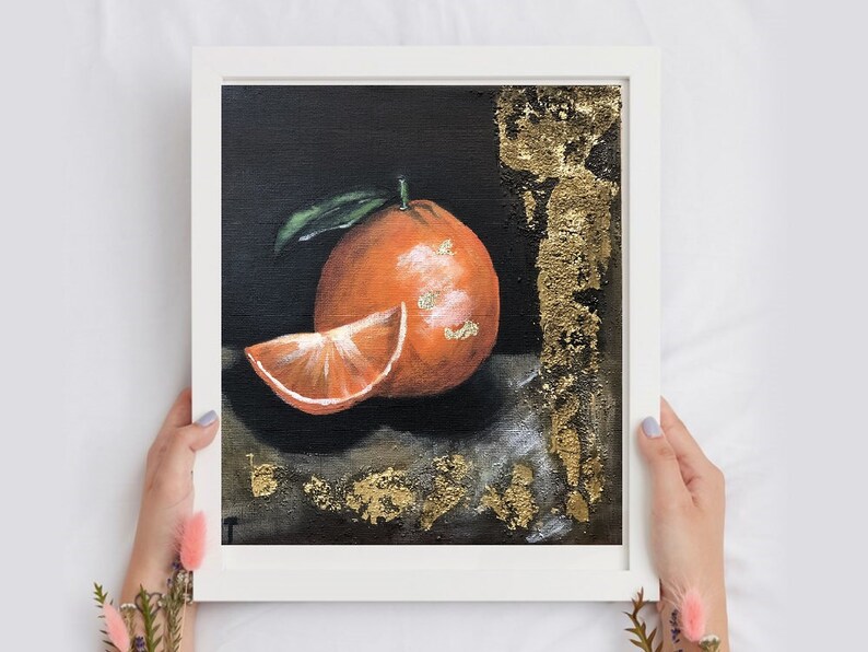 Puede incluir: Pintura de una naranja en un marco blanco. La obra de arte representa una naranja entera con una hoja verde y una rodaja de naranja. Acentos de pan de oro adornan el fondo oscuro. La obra de arte es sostenida por una persona.