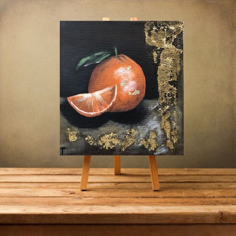 Puede incluir: Pintura de una naranja sobre un peque&ntilde;o caballete de madera. La obra de arte representa una naranja entera y una rodaja, con una hoja verde. Acentos de pan de oro adornan el fondo oscuro y los bordes, agregando textura y contraste a la naturaleza muerta.