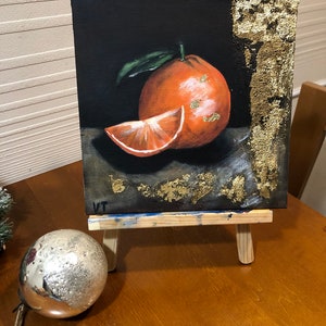 Puede incluir: Pintura de una naranja y una rodaja sobre un peque&ntilde;o lienzo, expuesta en un caballete de madera. La obra presenta una naranja entera con una hoja y una rodaja, sobre un fondo oscuro con detalles de pan de oro. Una bola plateada est&aacute; delante.