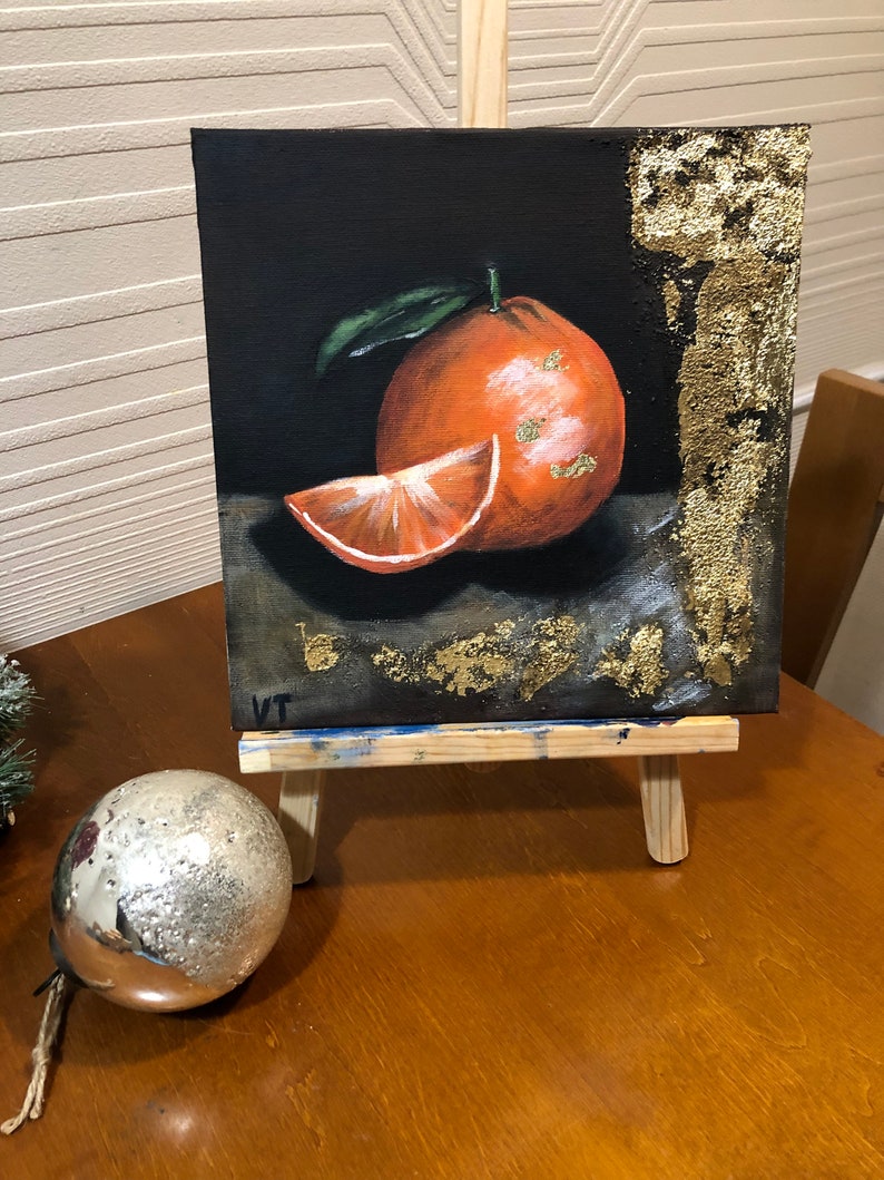 Puede incluir: Pintura de una naranja y una rodaja sobre un peque&ntilde;o lienzo, expuesta en un caballete de madera. La obra presenta una naranja entera con una hoja y una rodaja, sobre un fondo oscuro con detalles de pan de oro. Una bola plateada est&aacute; delante.