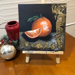 Puede incluir: Pintura acr&iacute;lica de una naranja y una rodaja sobre fondo negro, expuesta en un peque&ntilde;o caballete de madera. La naranja tiene una hoja verde y detalles dorados. Un portavelas rojo y una bola plateada est&aacute;n en el fondo.