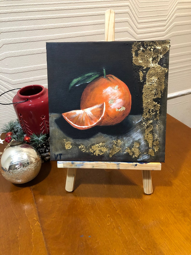 Puede incluir: Pintura acr&iacute;lica de una naranja y una rodaja sobre fondo negro, expuesta en un peque&ntilde;o caballete de madera. La naranja tiene una hoja verde y detalles dorados. Un portavelas rojo y una bola plateada est&aacute;n en el fondo.