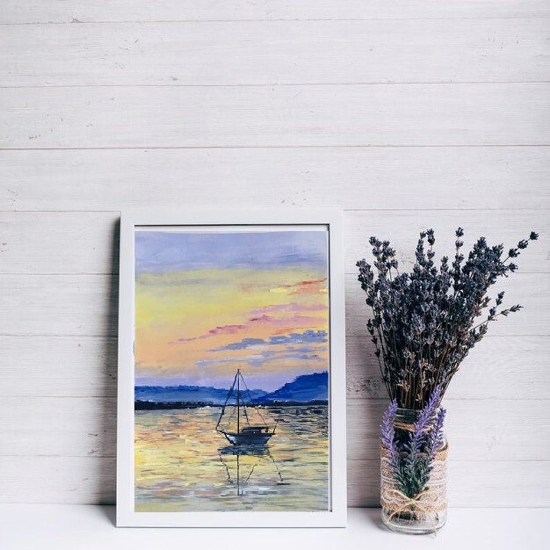 May include: A framed painting of a sailboat on the water at sunset. The artwork features a white frame, a colourful sky with yellow, orange, and blue hues, and a sailboat. A vase of lavender sits to the right.