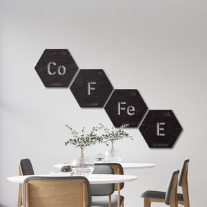 Puede incluir: Una mesa blanca con cuatro sillas grises se encuentra en una habitación con una pared blanca. La pared tiene una decoración de cinco letreros hexagonales de metal con los símbolos químicos y los nombres de cobalto, flúor, hierro y erbio.