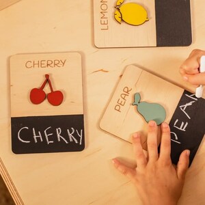 May include: A child is learning to spell fruit names using wooden blocks with chalkboard surfaces. The blocks feature images of a lemon, cherry, and pear. The child is writing the word "pear" on the chalkboard surface of the block.