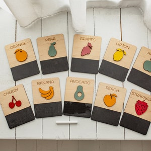 May include: A set of 12 wooden flashcards featuring different fruits and vegetables. Each card has a colorful illustration of the fruit or vegetable and the name printed on the top half. The bottom half of each card is a black chalkboard surface.