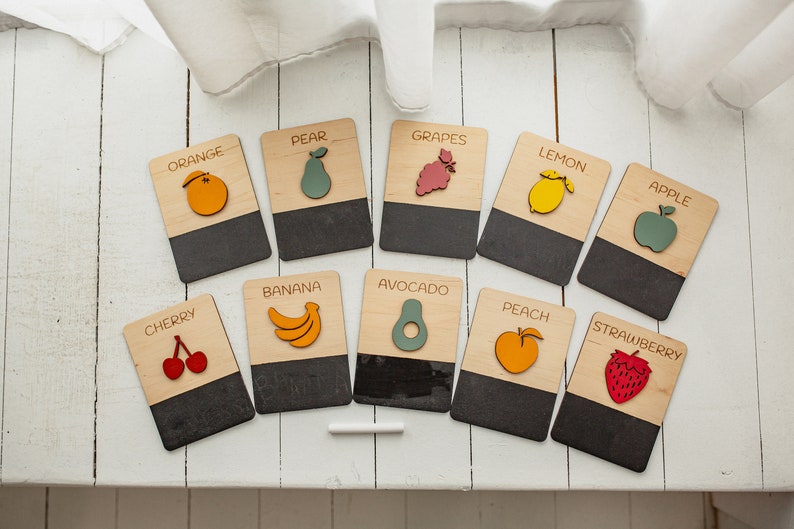 May include: A set of 12 wooden flashcards featuring different fruits and vegetables. Each card has a colorful illustration of the fruit or vegetable and the name printed on the top half. The bottom half of each card is a black chalkboard surface.