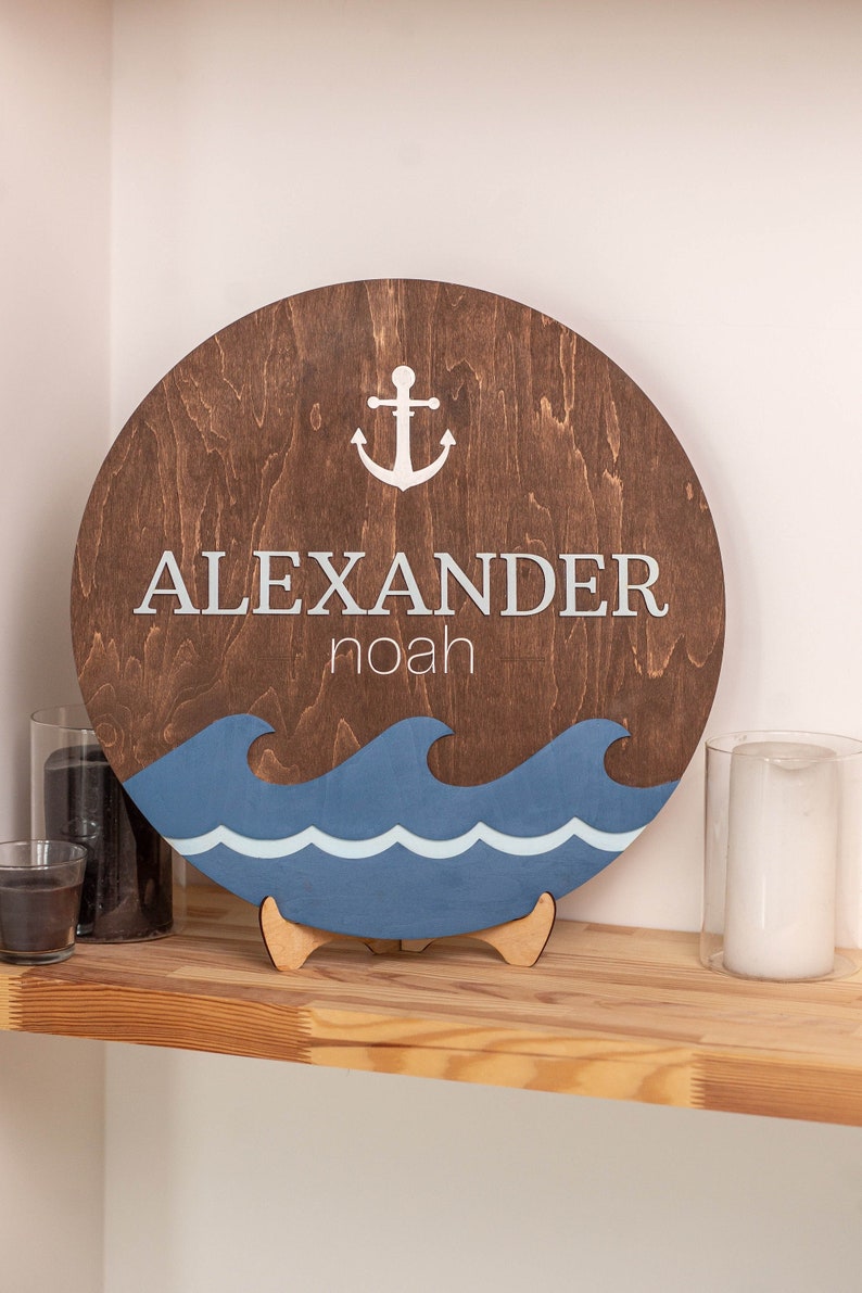 May include: A round wooden sign with a blue and white wave design at the bottom. The sign has a white anchor and the text "ALEXANDER noah" in white.