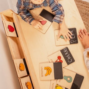May include: A wooden playset with a child playing with colorful wooden fruit cards. The cards have the name of the fruit written on them, such as "banana", "pear", "cherry", and "lemon".