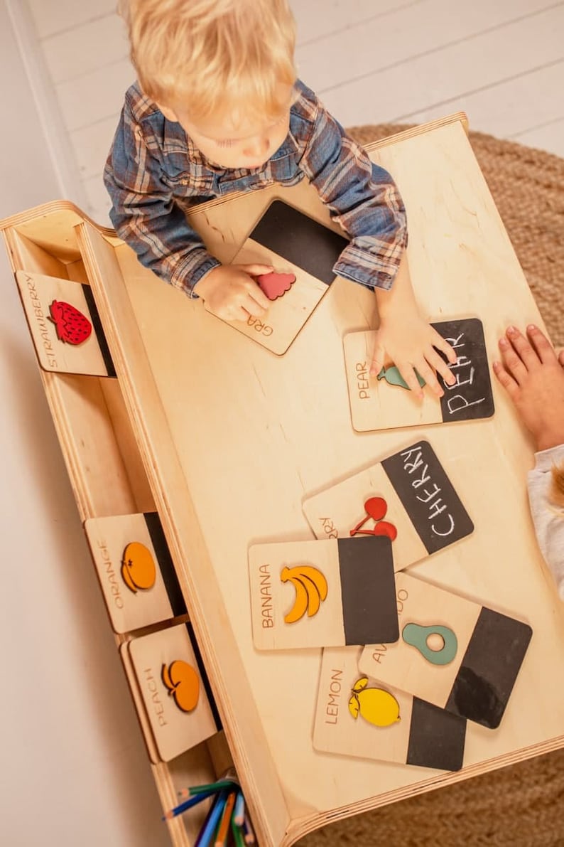 May include: A wooden playset with a child playing with colorful wooden fruit cards. The cards have the name of the fruit written on them, such as "banana", "pear", "cherry", and "lemon".
