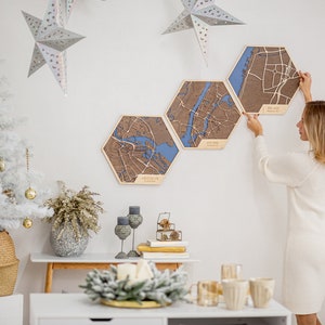May include: Three hexagonal wooden wall maps with blue and brown details. The maps are hanging on a white wall. A woman is hanging the maps on the wall. The maps are of different cities. The cities are labeled on the maps. The maps are a unique and stylish way to decorate a home.