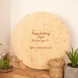May include: A round wooden plaque with a heart-shaped stand. The plaque has a personalized message that reads "Happy birthday, Kylie! We love you. Your mom and dad."