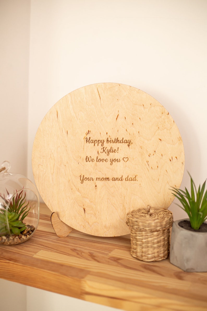May include: A round wooden plaque with a heart-shaped stand. The plaque has a personalized message that reads "Happy birthday, Kylie! We love you. Your mom and dad."