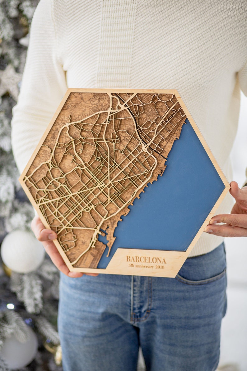 May include: A wooden map of Barcelona with a blue background. The map is engraved with the city's streets and landmarks. The text "Barcelona 35th anniversary 2018" is printed on the bottom of the map.