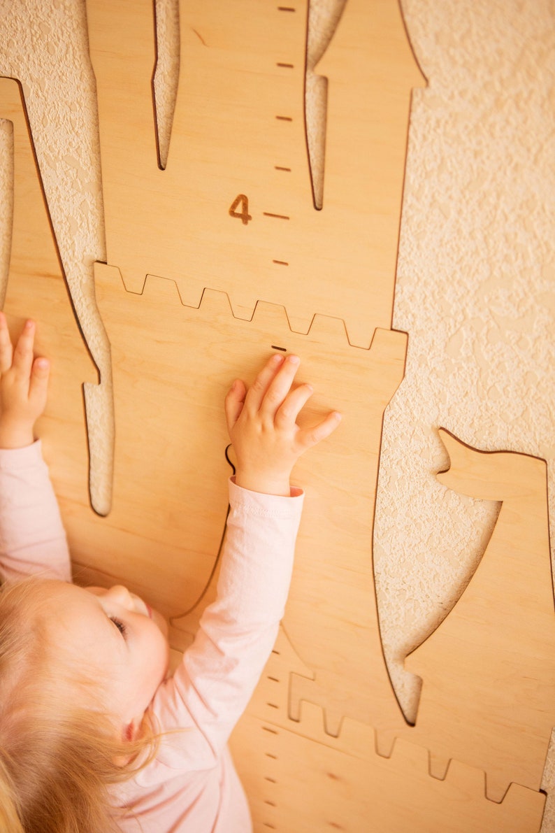 May include: A child reaches up to a wooden castle-shaped growth chart with a number "4" marked on it. The chart is mounted on a wall.