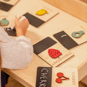 May include: A wooden learning toy with black chalkboard cards featuring different fruits. The cards have the name of the fruit written on them, and a wooden cutout of the fruit. The fruits include a pear, lemon, strawberry, and cherry.