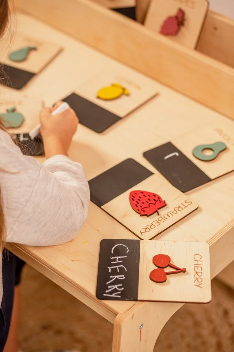 May include: A wooden learning toy with black chalkboard cards featuring different fruits. The cards have the name of the fruit written on them, and a wooden cutout of the fruit. The fruits include a pear, lemon, strawberry, and cherry.