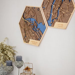 May include: Two hexagonal wooden wall decor pieces featuring laser-cut maps of Amsterdam and New York City. The maps are brown with gold accents and have a blue background. The Amsterdam map has the text "AMSTERDAM Weekend 2016" at the bottom.
