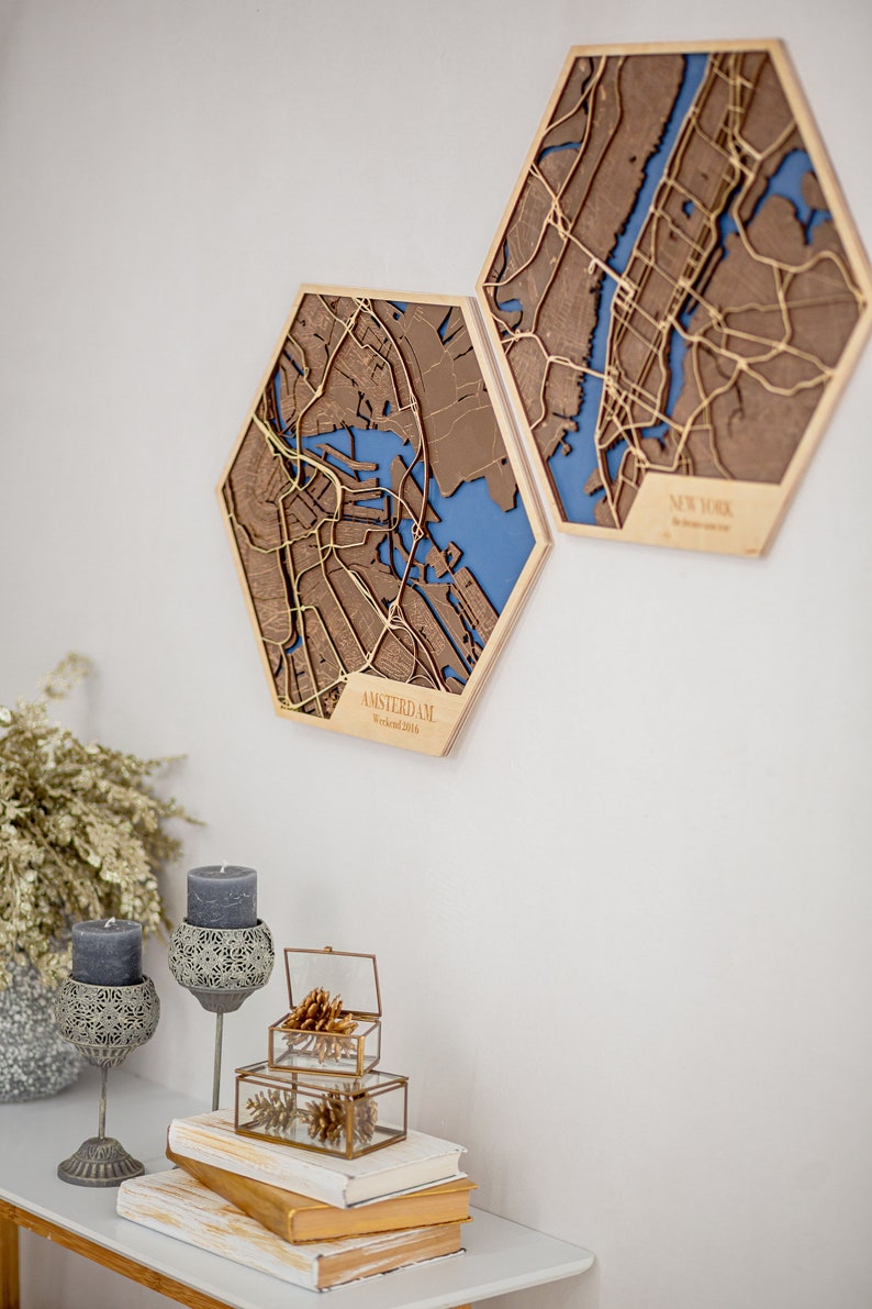 May include: Two hexagonal wooden wall decor pieces featuring laser-cut maps of Amsterdam and New York City. The maps are brown with gold accents and have a blue background. The Amsterdam map has the text "AMSTERDAM Weekend 2016" at the bottom.