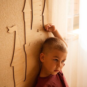 May include: A young child stands next to a wooden castle-shaped height chart, reaching up to the number "4" on the ruler. The child is wearing a red shirt and has short blond hair.
