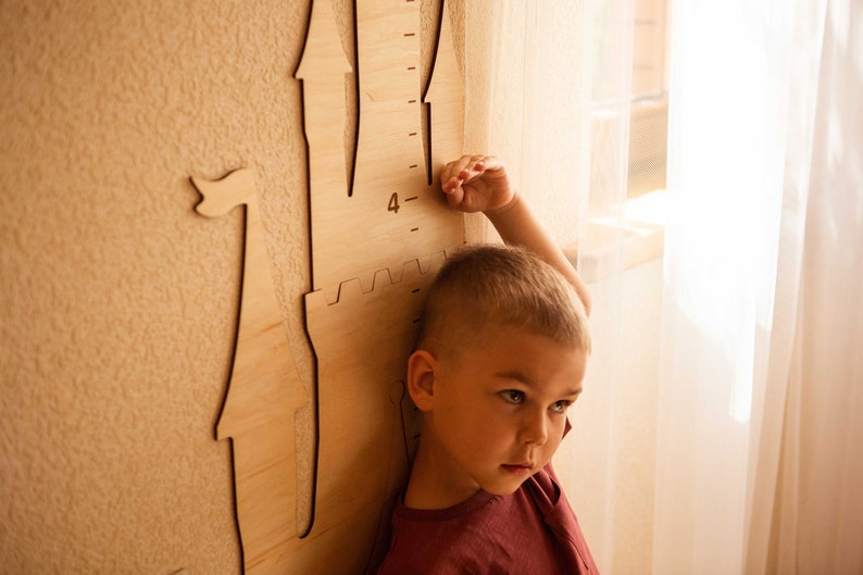May include: A young child stands next to a wooden castle-shaped height chart, reaching up to the number "4" on the ruler. The child is wearing a red shirt and has short blond hair.