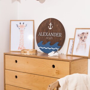 May include: A wooden round sign with the name "ALEXANDER noah" and an anchor symbol. The sign is painted in brown, blue, and white colors. The sign is hanging on a wall with a white background. There are also other decorations on the wall, including a stuffed rhinoceros head and a string of flags.