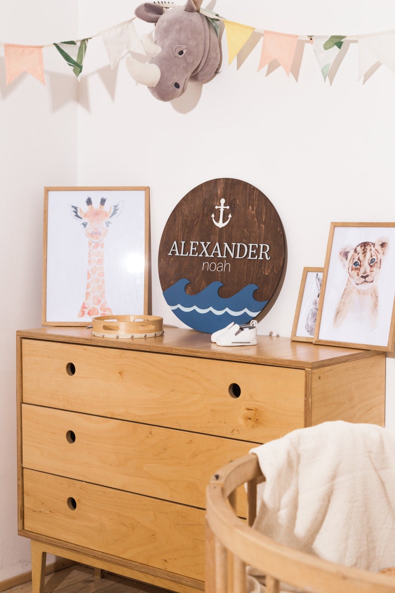 May include: A wooden round sign with the name "ALEXANDER noah" and an anchor symbol. The sign is painted in brown, blue, and white colors. The sign is hanging on a wall with a white background. There are also other decorations on the wall, including a stuffed rhinoceros head and a string of flags.