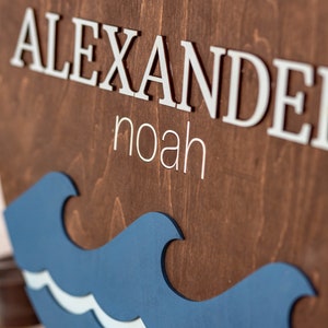 May include: A wooden sign with the name "Alexander Noah" in white letters. The sign is decorated with a blue and white wave design and an anchor.