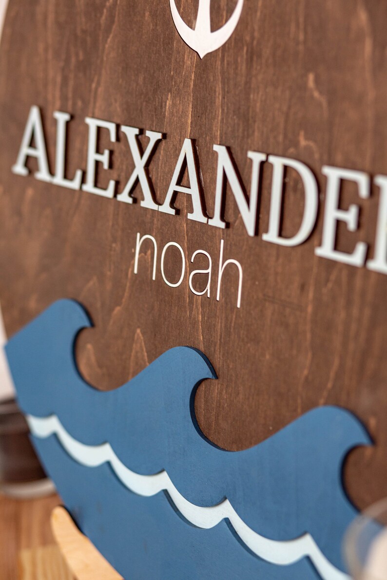May include: A wooden sign with the name "Alexander Noah" in white letters. The sign is decorated with a blue and white wave design and an anchor.