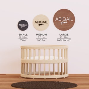 May include: Three wooden circles with the name "Abigail grace" written on them in black lettering. The circles are in three different sizes: small (11.5 inches), medium (17 inches), and large (23 inches). The small circle is black, the medium circle is natural wood, and the large circle is dark walnut. The circles are hanging on a white wall.