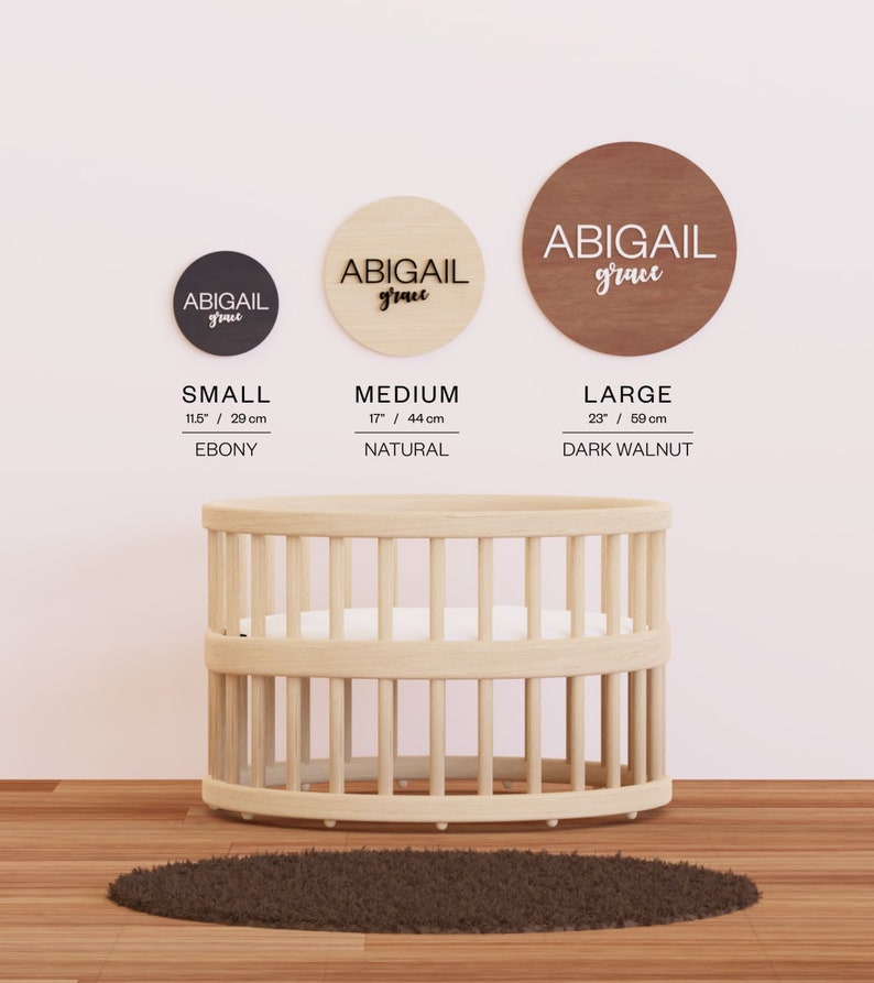 May include: Three wooden circles with the name "Abigail grace" written on them in black lettering. The circles are in three different sizes: small (11.5 inches), medium (17 inches), and large (23 inches). The small circle is black, the medium circle is natural wood, and the large circle is dark walnut. The circles are hanging on a white wall.